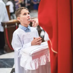 Chłopiec w albie przyjmuje komunię świętą na tle kościelnego wnętrza, z widocznym detalem haftowanego emblematu na stroju.