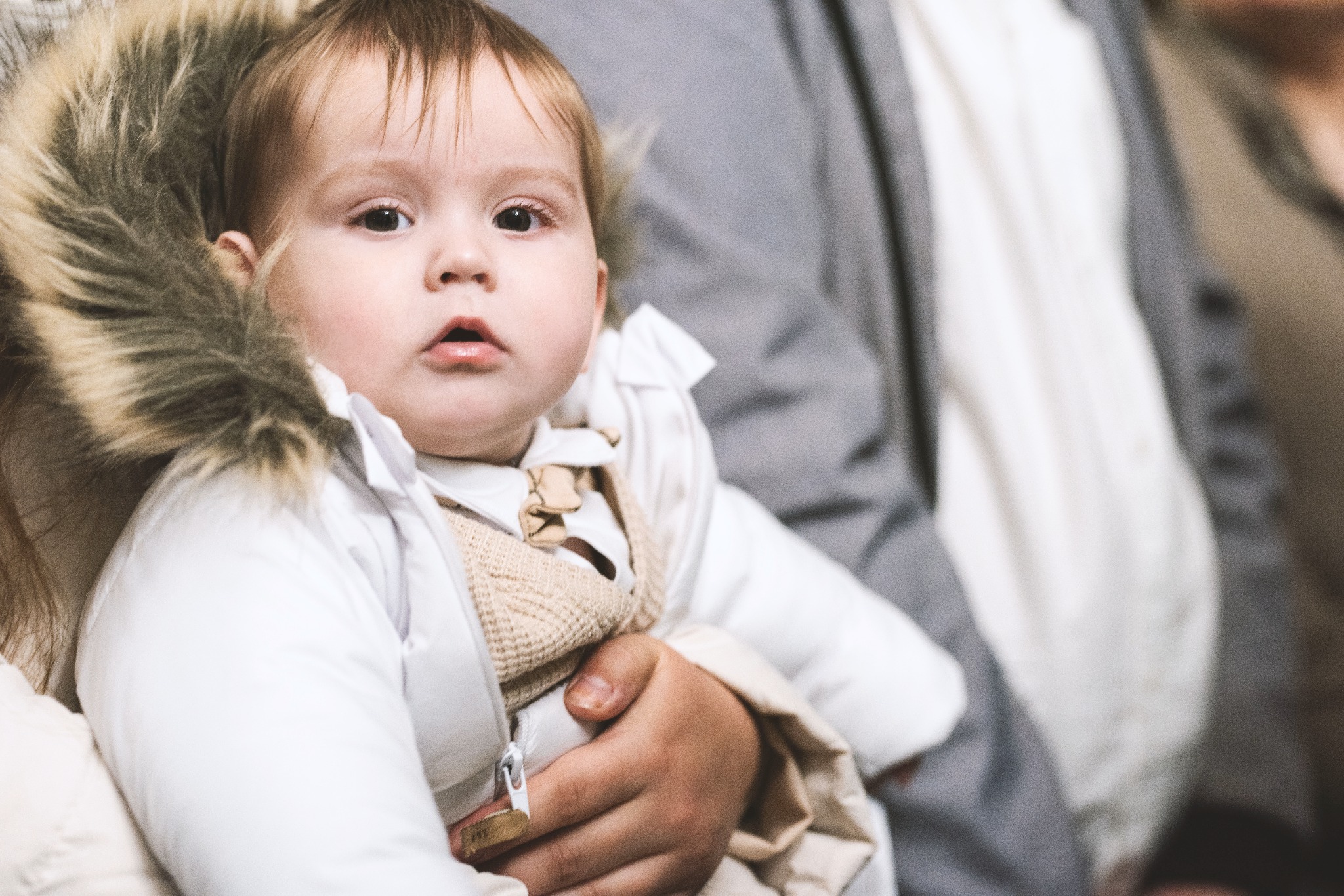 Portret niemowlęcia w białej kurtce z futrzanym kapturem, trzymanego na rękach przez osobę dorosłą, z neutralnym wyrazem twarzy i miękkim oświetleniem.