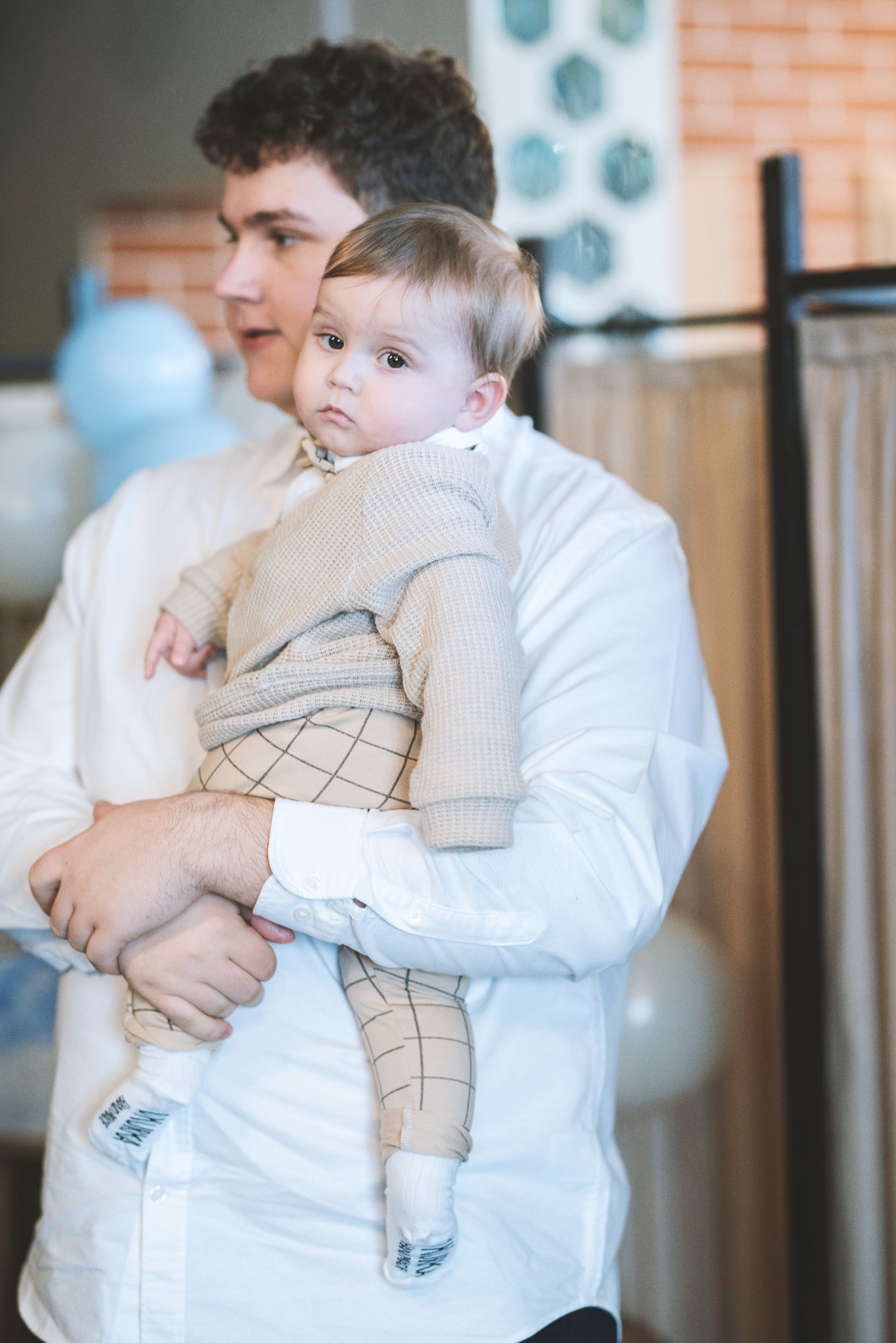 Mężczyzna w białej koszuli trzyma na rękach niemowlę ubrane w sweterek i spodnie w kratę, widoczne skarpetki z napisem.