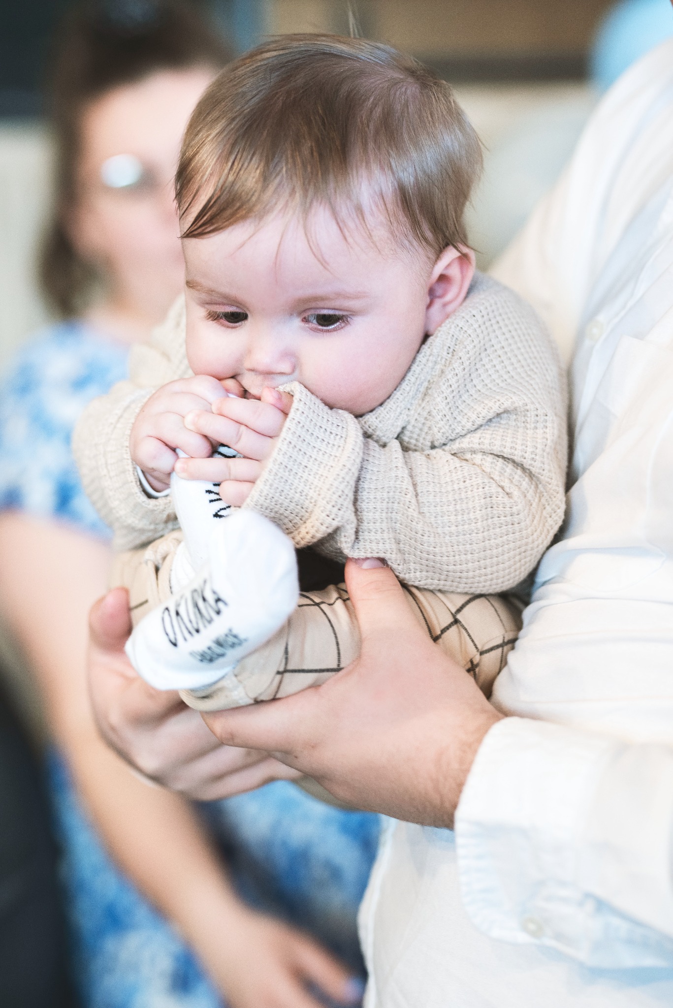 Niemowlę w beżowym swetrze i spodniach w kratkę trzymane na rękach, ssie białą skarpetkę z napisem 'OKOKKA' na tle rozmytej postaci kobiety.