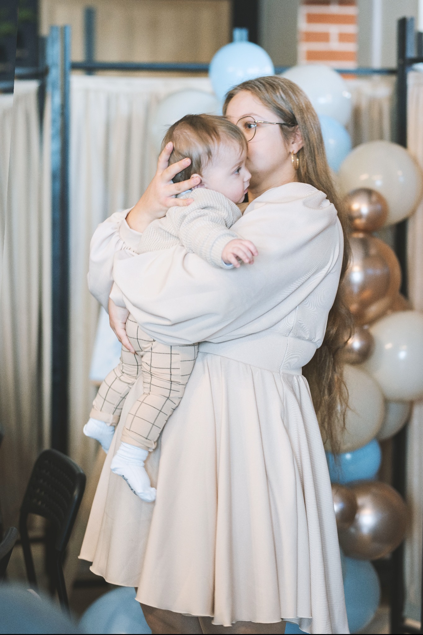 Matka w beżowej sukience i okularach przytula małego synka ubranego w sweterek i spodnie w kratę na tle dekoracji z balonów w odcieniach błękitu i złota.