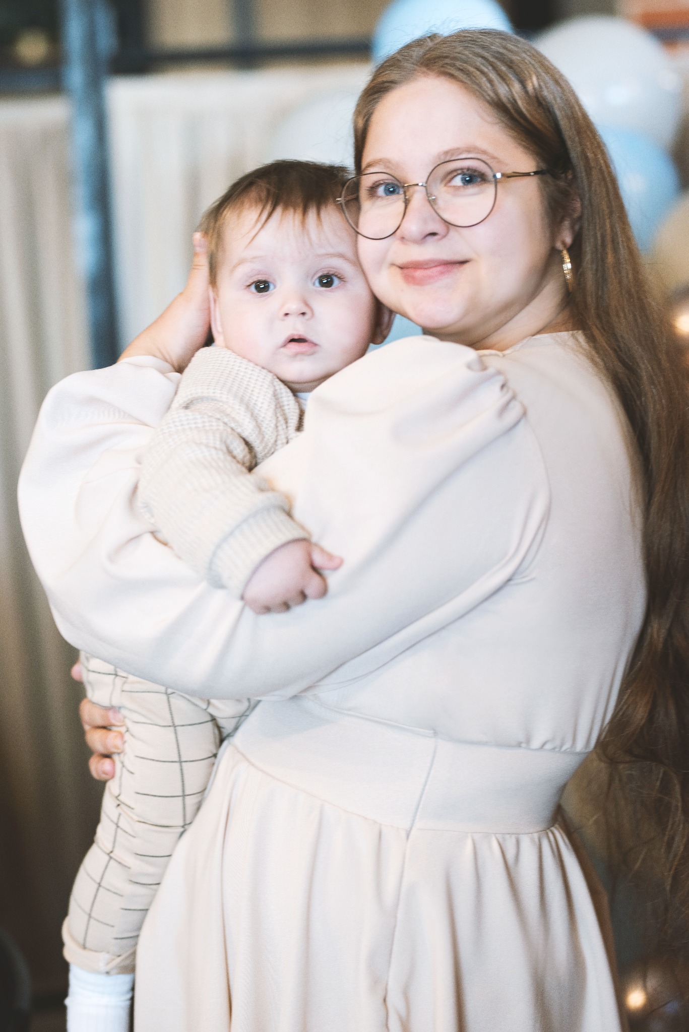 Uśmiechnięta kobieta w okularach obejmuje małe dziecko ubrane w sweterek i spodnie w kratę na tle jasnych balonów.