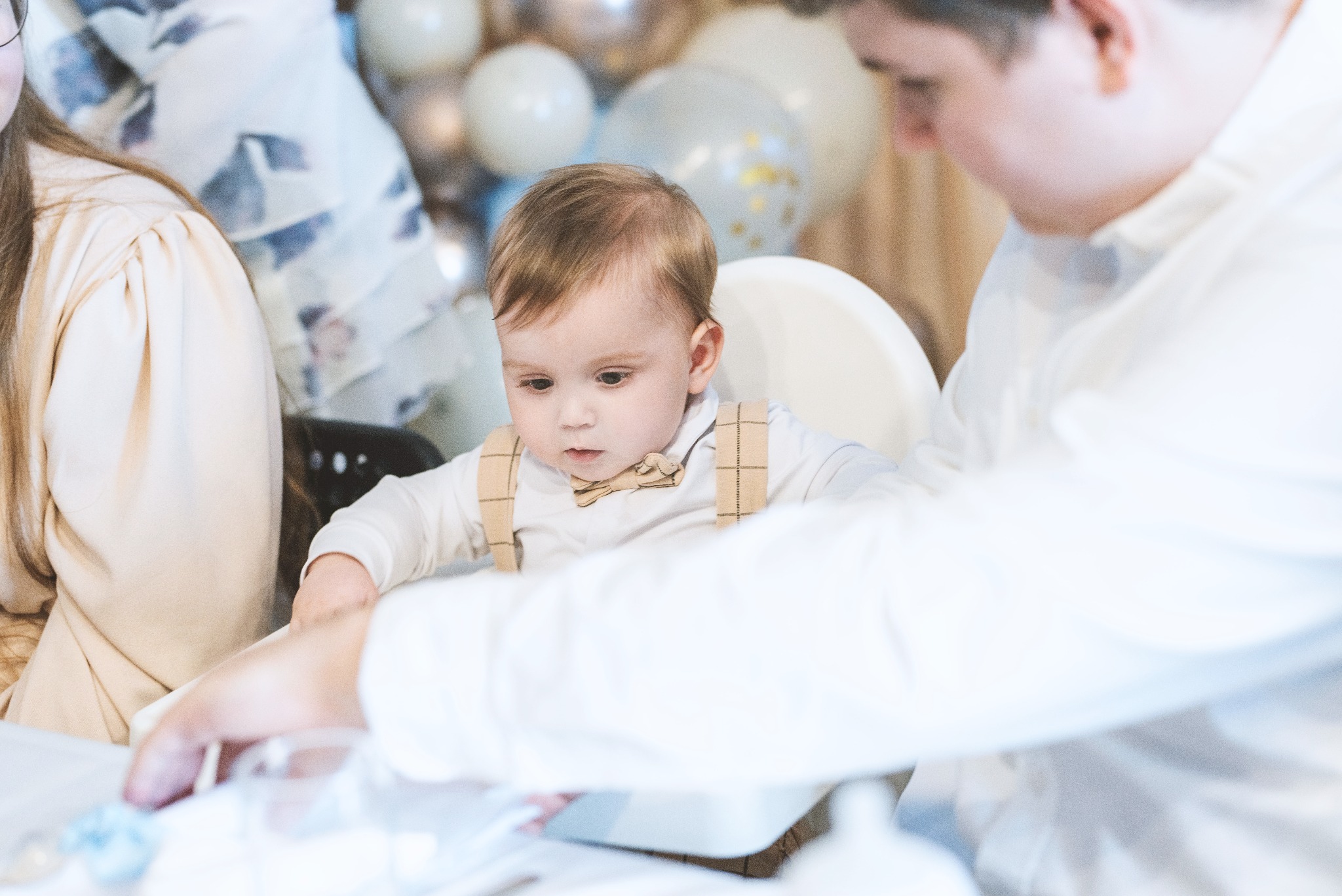 Mały chłopiec w muszce i szelkach siedzi w krzesełku do karmienia, otoczony dekoracjami z jasnych balonów podczas uroczystości.