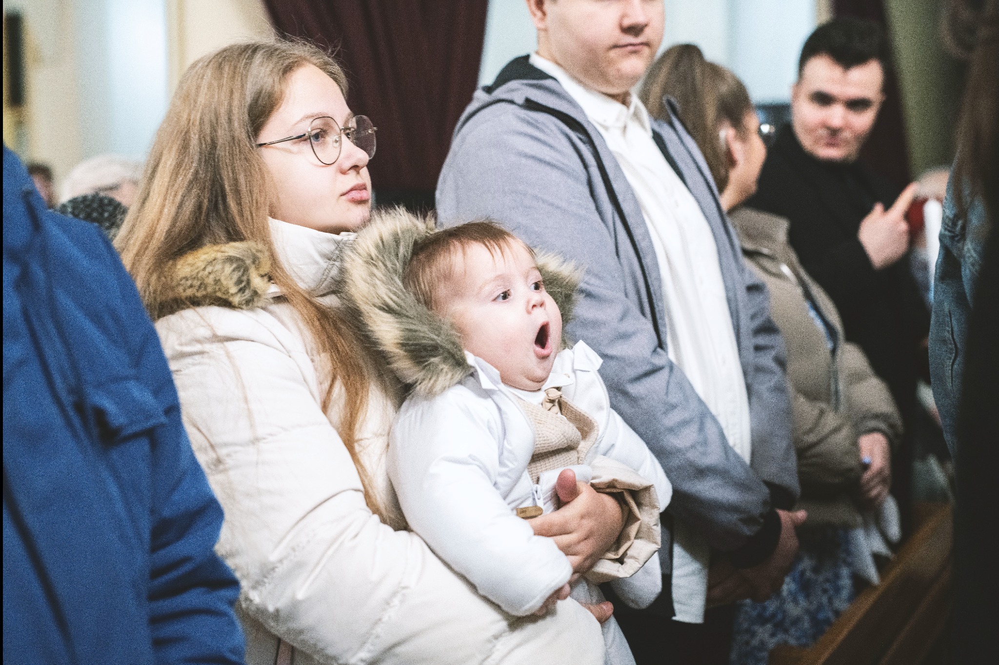 Młoda kobieta w okularach trzyma na rękach szeroko otwierającego buzię niemowlaka w białym kombinezonie z futrzanym kapturem, w tle widoczni inni ludzie, prawdopodobnie w kościele.