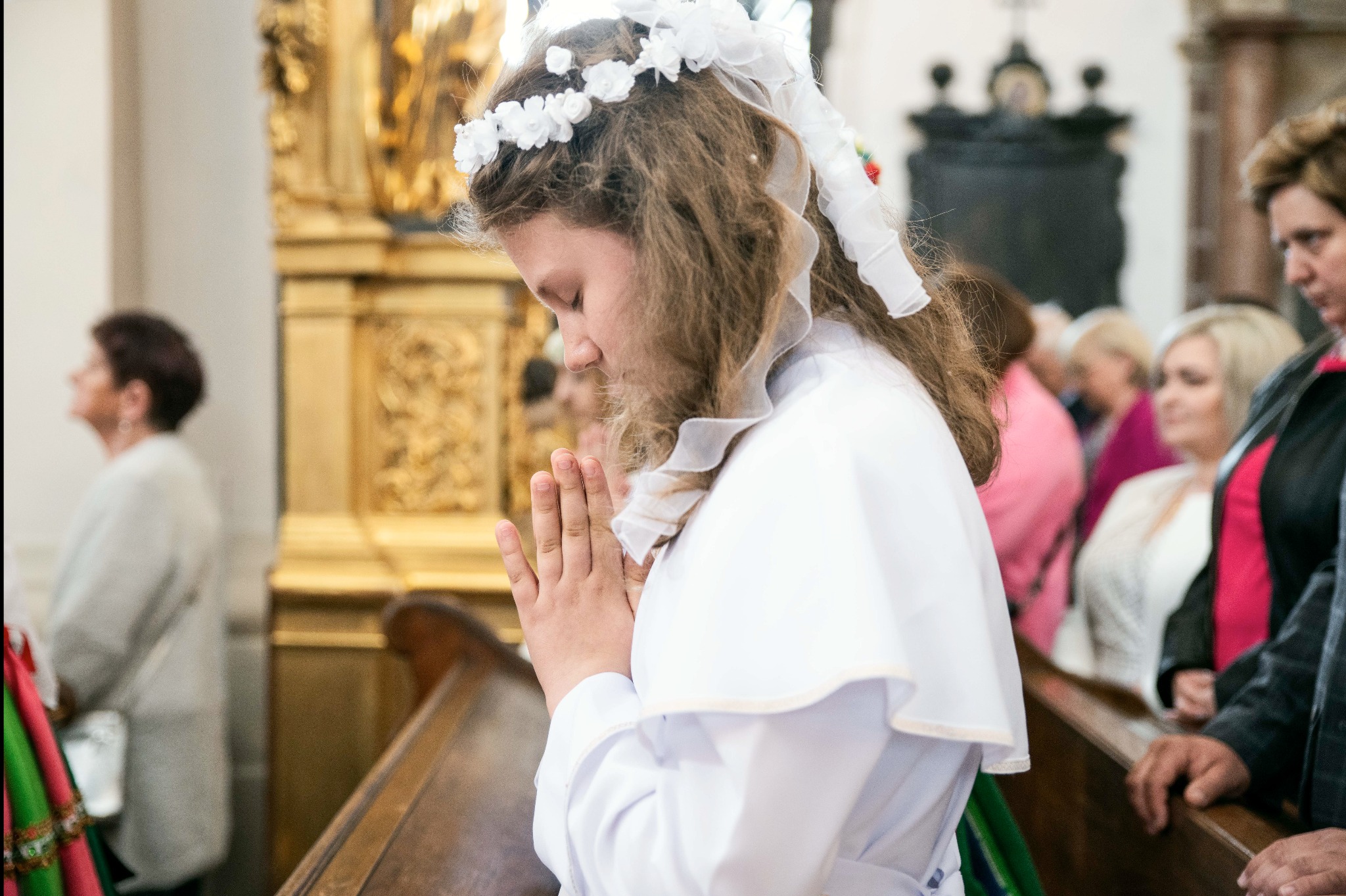 Dziewczynka w białej albie i wianku klęczy w ławce kościelnej, modląc się podczas ceremonii Pierwszej Komunii Świętej, z rękami złożonymi do modlitwy.