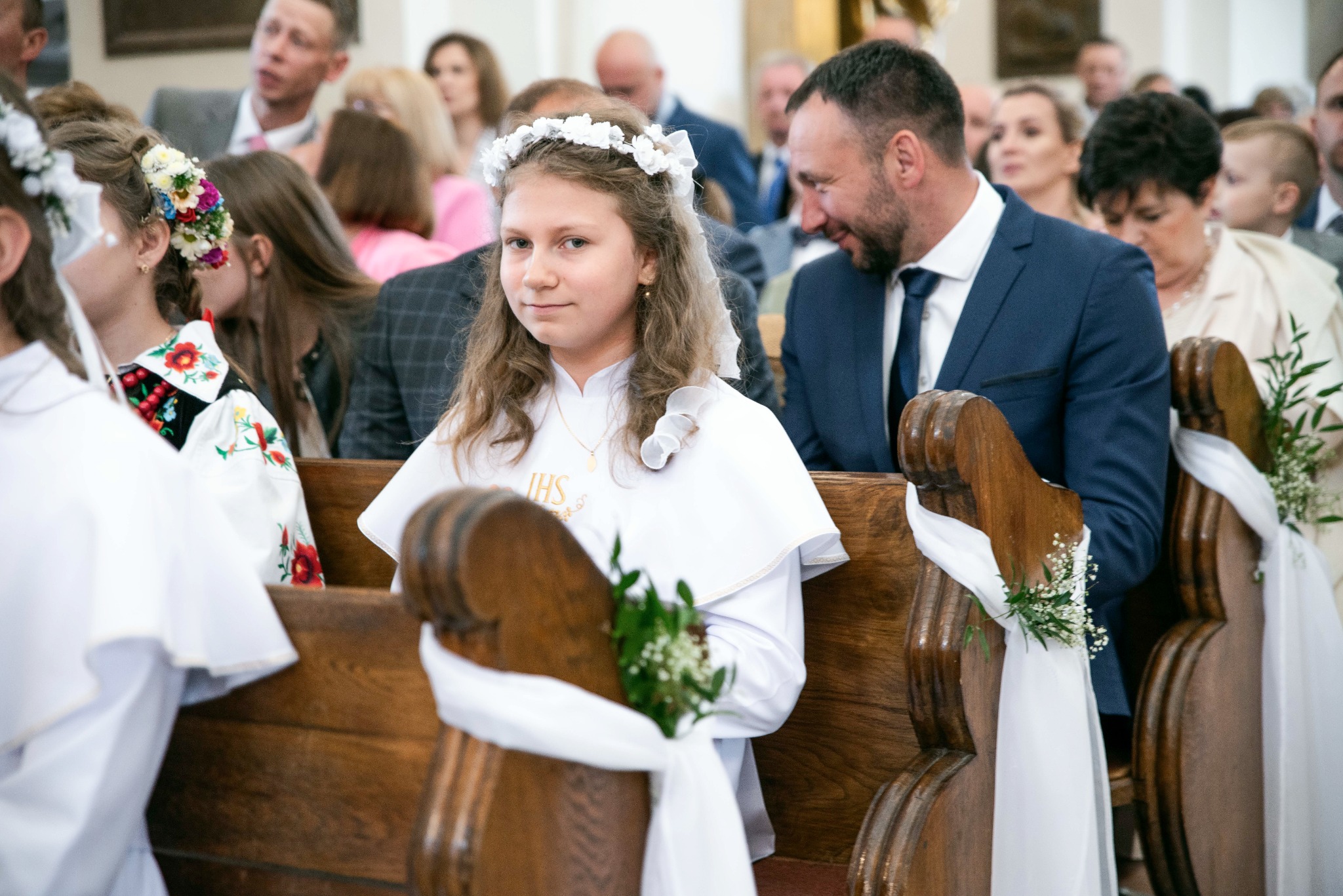 Dziewczynka w białej albie z wiankiem na głowie siedzi w ławce kościelnej, udekorowanej białymi wstążkami i kwiatami, podczas uroczystości Pierwszej Komunii Świętej.