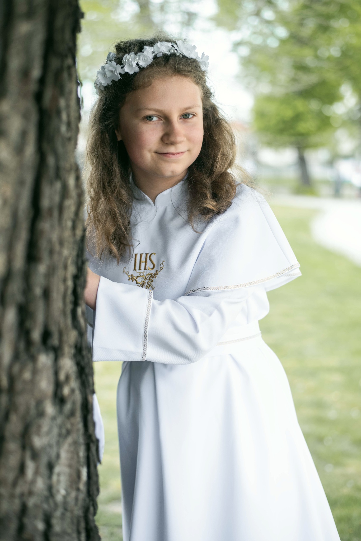 Portret dziewczynki w białej albie komunijnej, z wiankiem na głowie, opierającej się o korę drzewa na tle rozmytej zieleni.