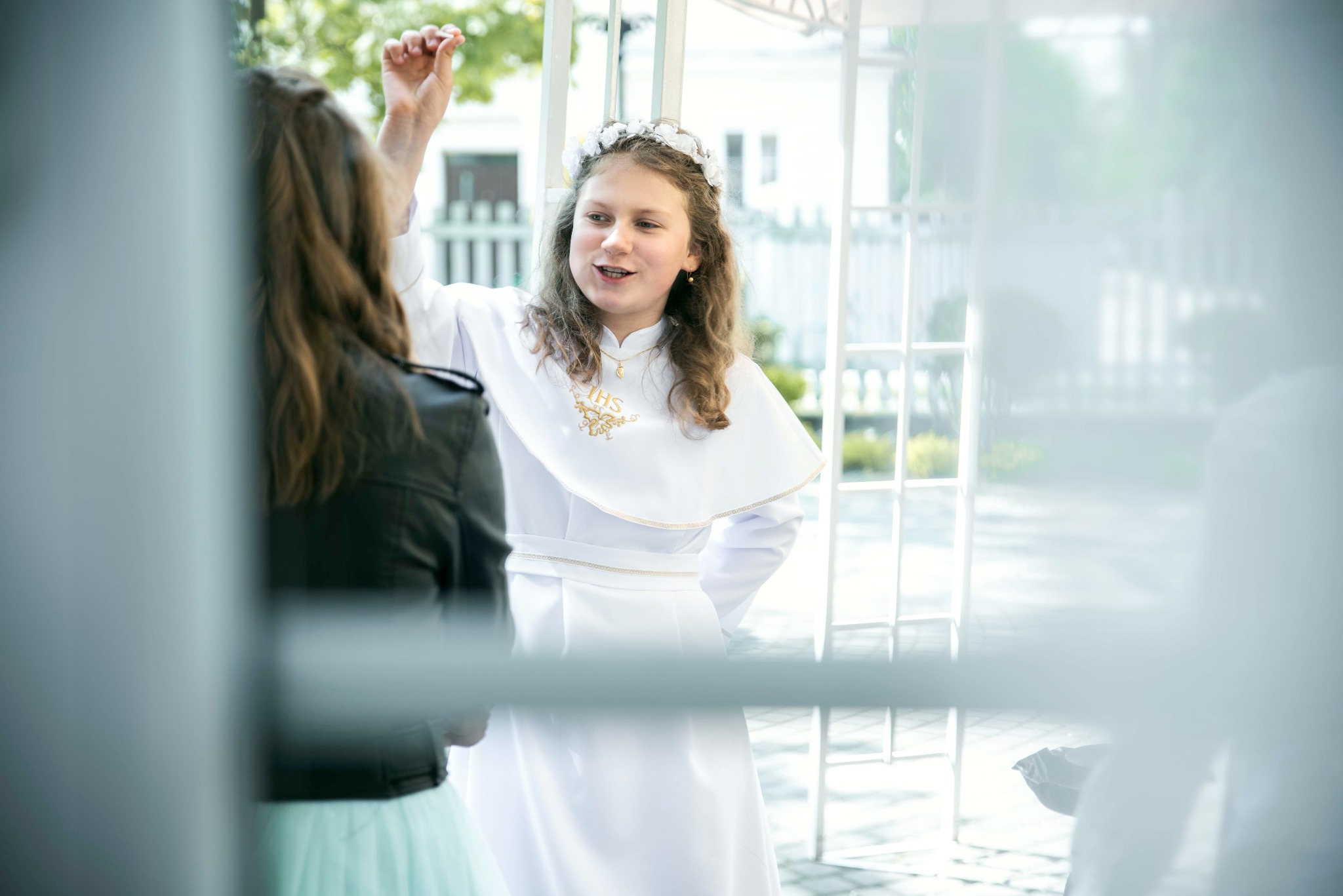 Dziewczynka w białej albie komunijnej z wiankiem na głowie, rozmawia z osobą w czarnej kurtce i turkusowej spódnicy na tle białej konstrukcji.
