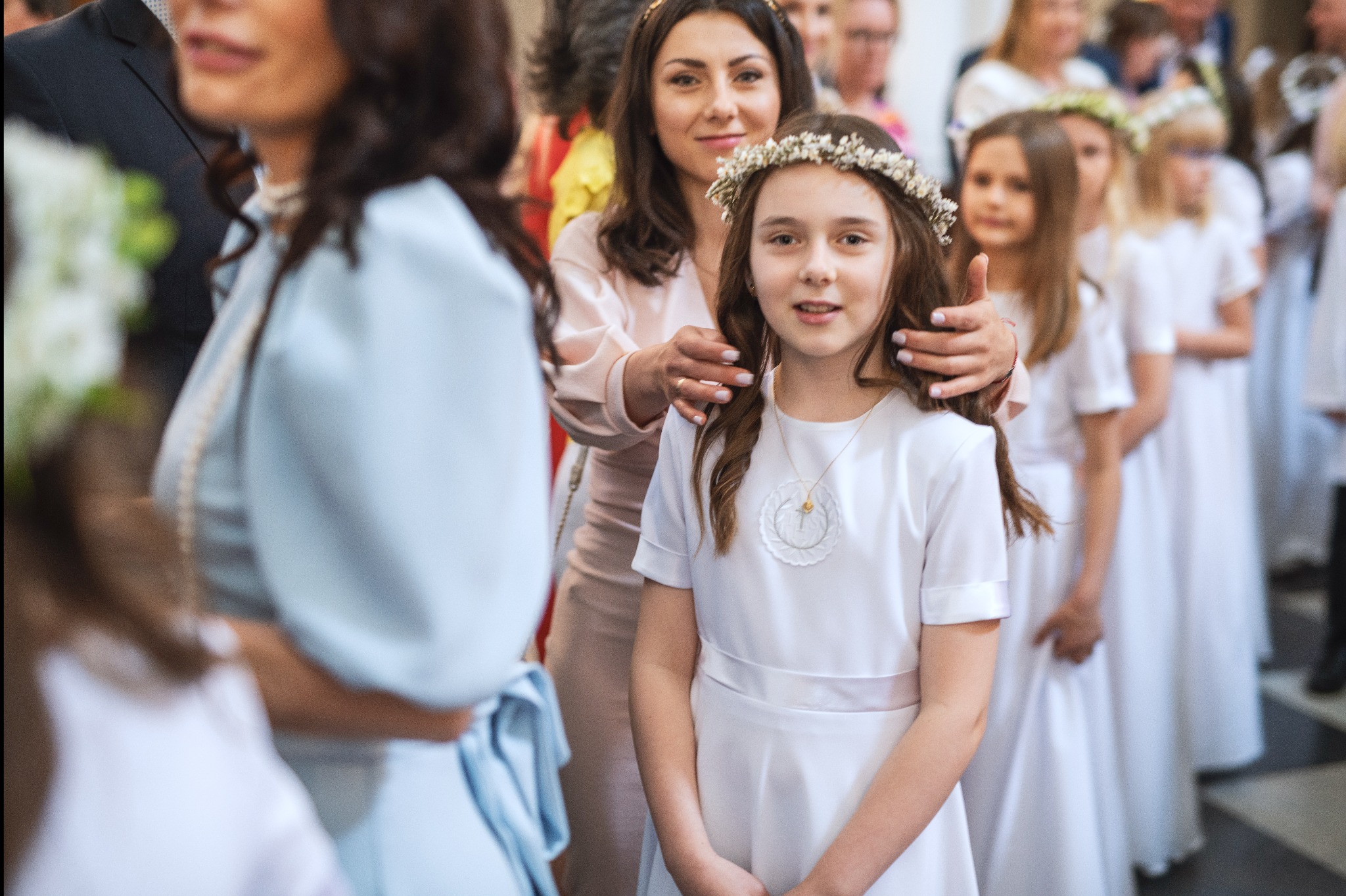 Dziewczynka w białej sukience komunijnej z wiankiem na głowie, wspierana przez matkę, w tle rząd innych dzieci w podobnych strojach.