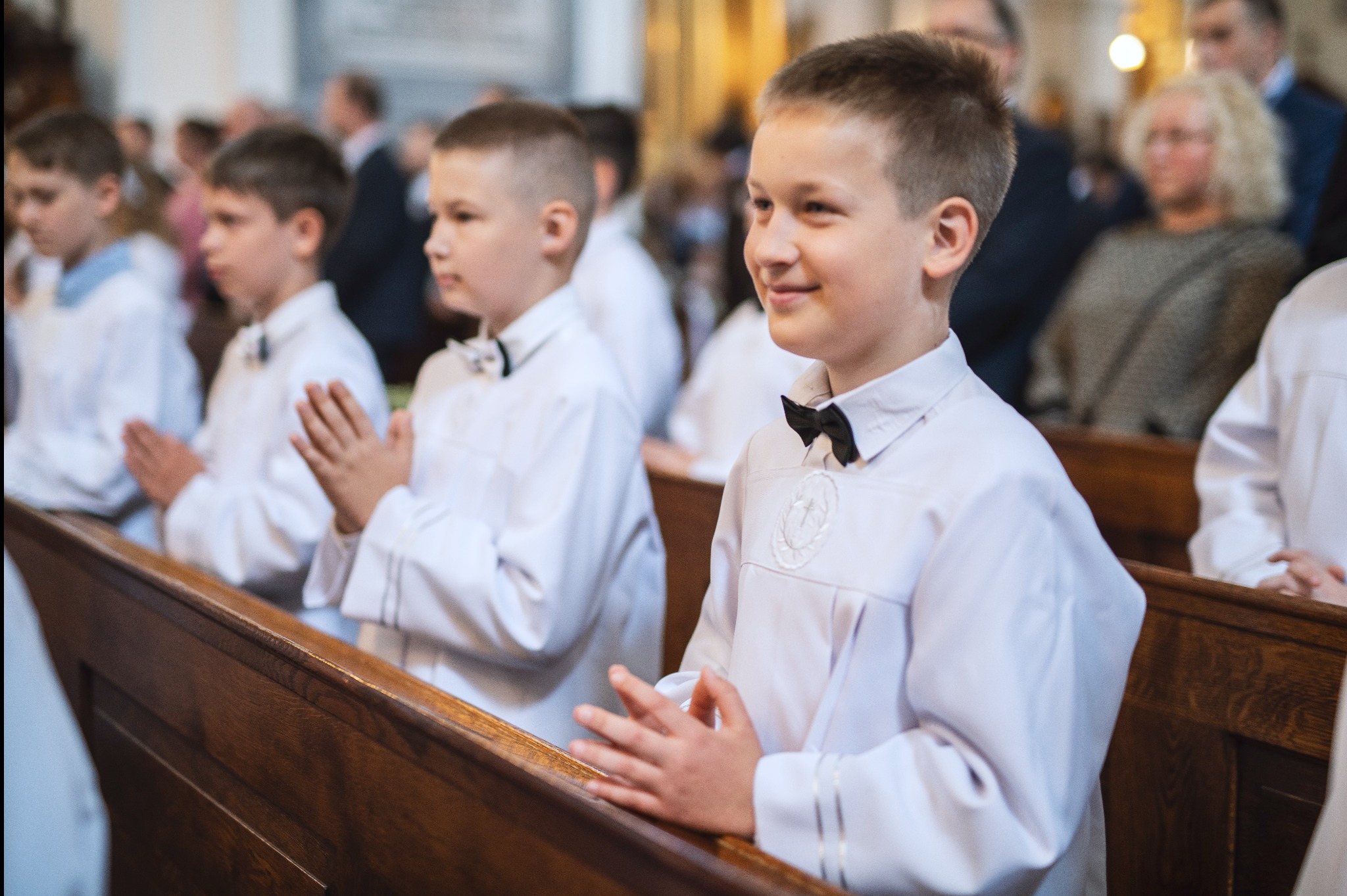 Chłopcy w białych albach i muchach, siedzący w ławkach kościelnych podczas uroczystości religijnej, z uśmiechniętym chłopcem na pierwszym planie.