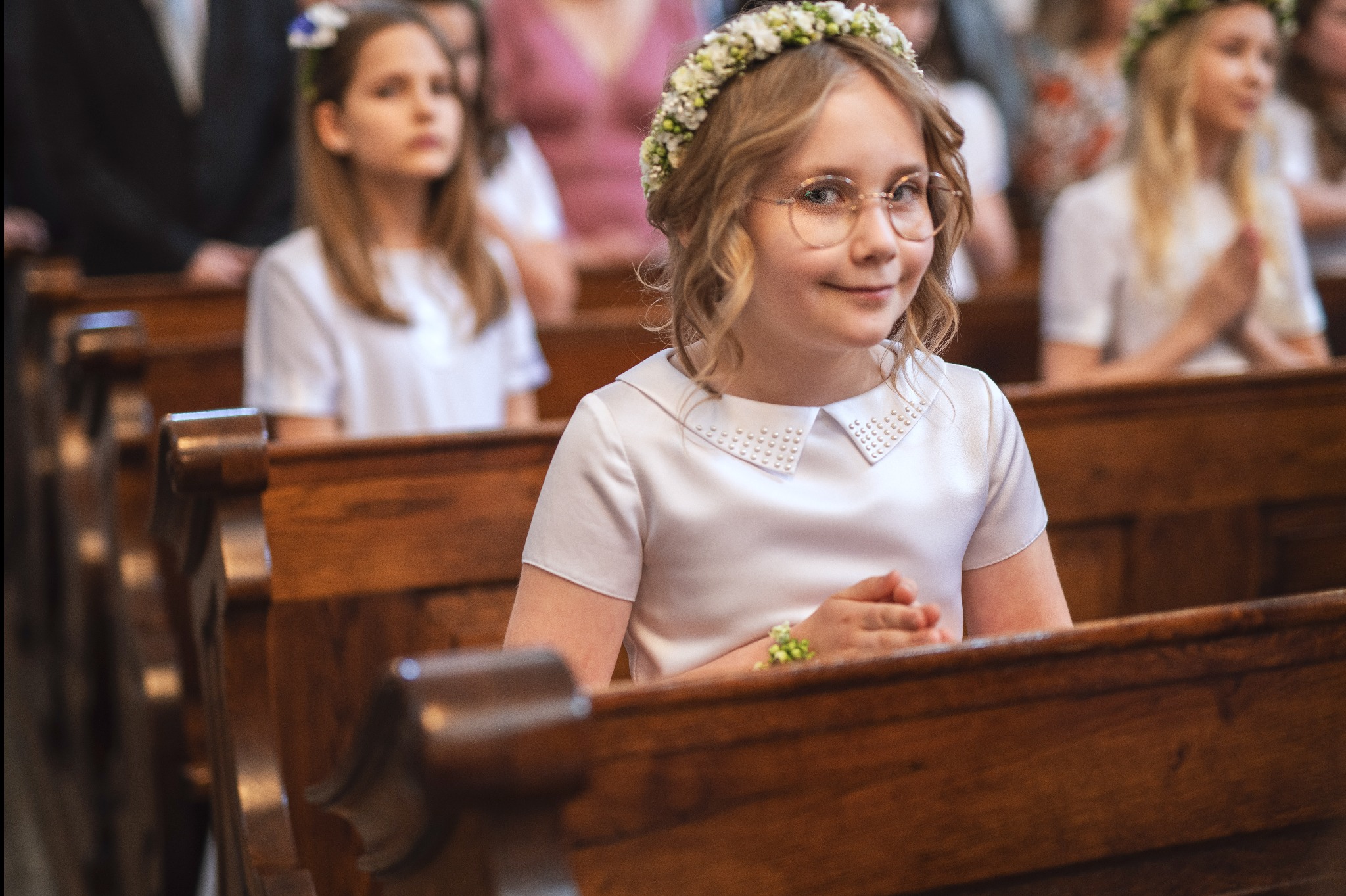 Dziewczynka w okularach i wianku z białych kwiatów, siedząca w ławce kościelnej podczas uroczystości, z delikatną ozdobą kwiatową na nadgarstku.