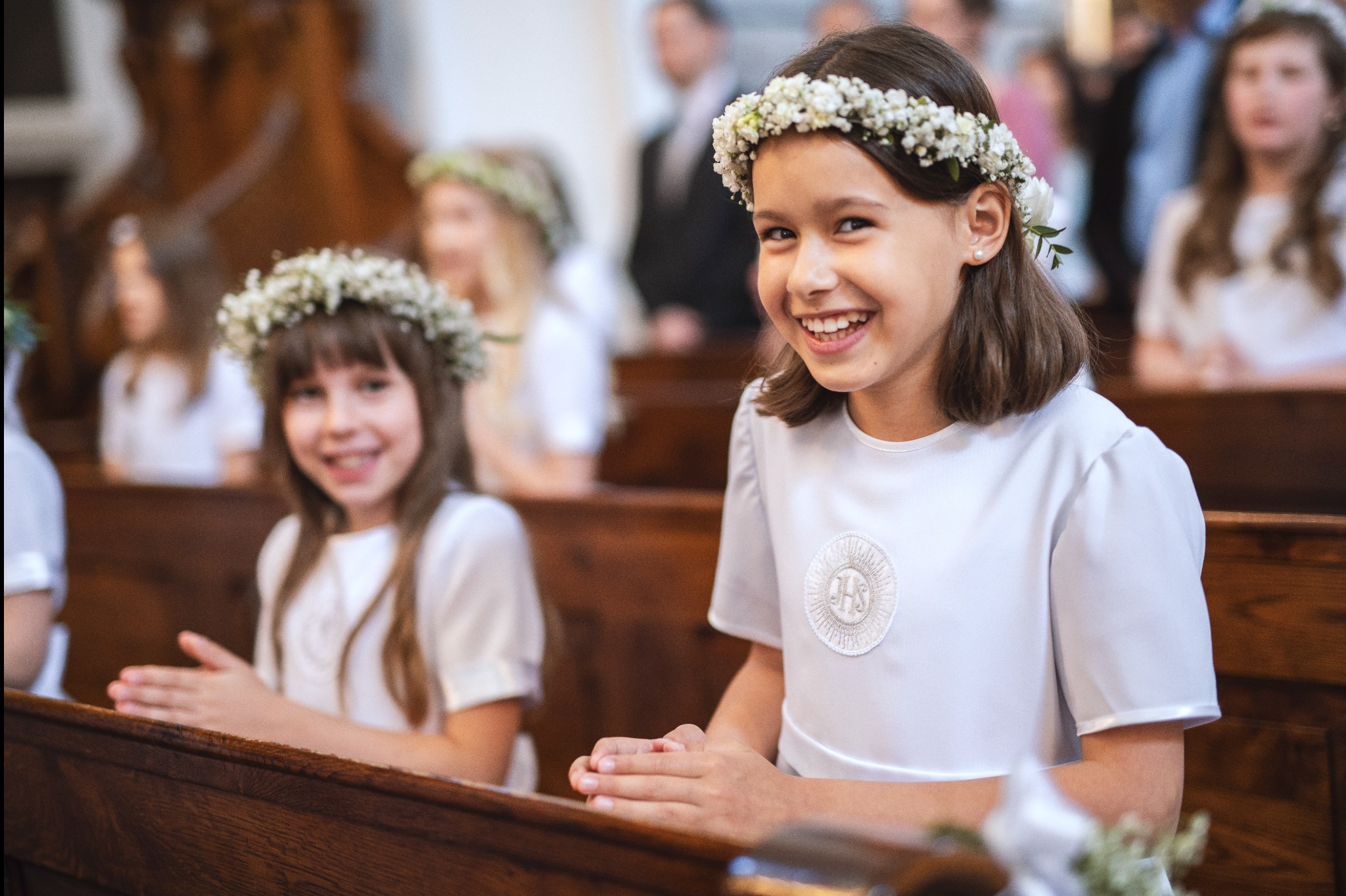 Dziewczynka w białej albie i wianku z białych kwiatów uśmiecha się siedząc w ławce kościoła podczas uroczystości religijnej, w tle inne dzieci również w wiankach.