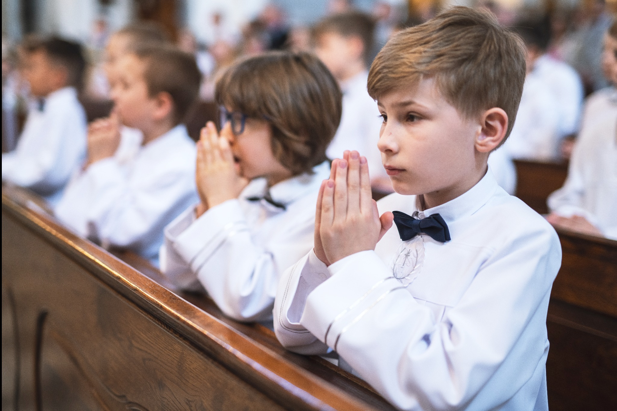 Chłopcy w białych strojach komunijnych, w ławkach kościelnych, z rękami złożonymi do modlitwy, podczas uroczystości religijnej.