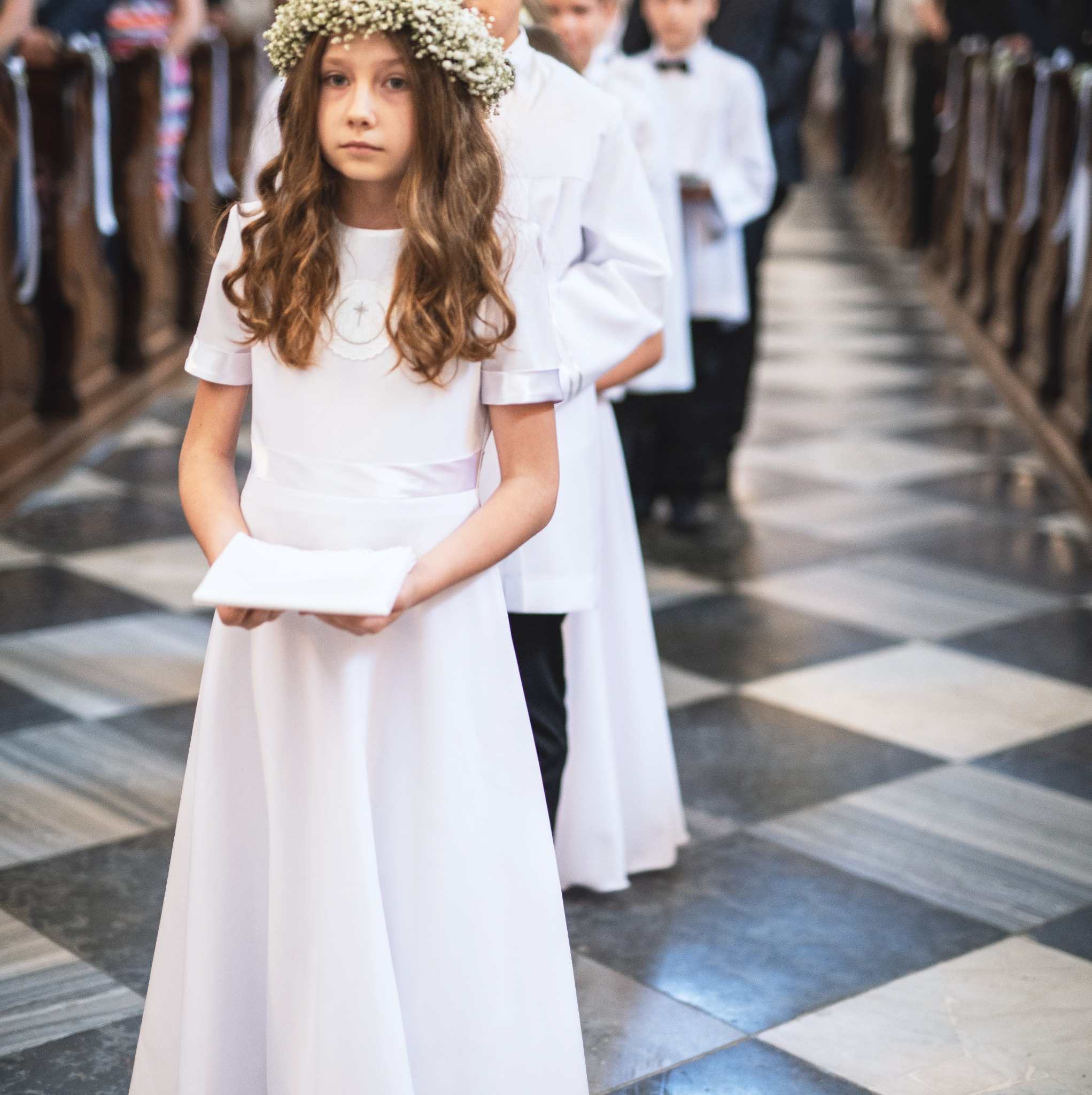 Dziewczynka w białej albie i wianku z białych kwiatów, trzymająca kartkę, stoi w rzędzie z innymi dziećmi podczas uroczystości w kościele z szachownicową posadzką.