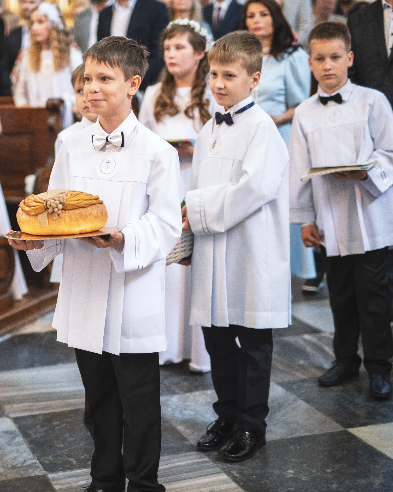 Chłopiec w białej albie i z muszką niesie udekorowany chleb na złotej tacy podczas uroczystości religijnej w kościele.