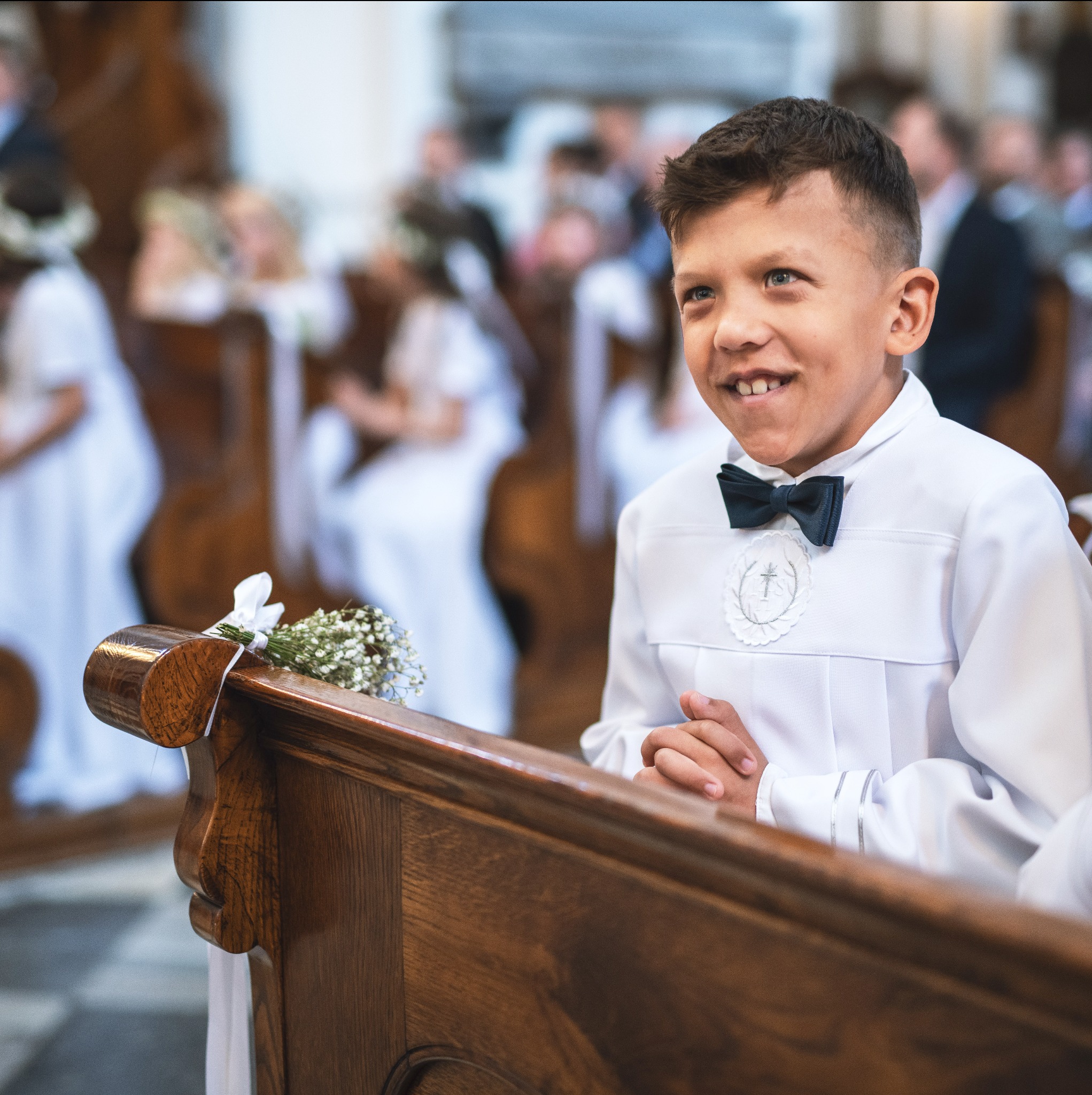 Chłopiec w stroju komunijnym z muchą, siedzący w ławce kościelnej, z bukiecikiem kwiatów na poręczy i uśmiechem na twarzy, w tle inne dzieci ubrane na biało.
