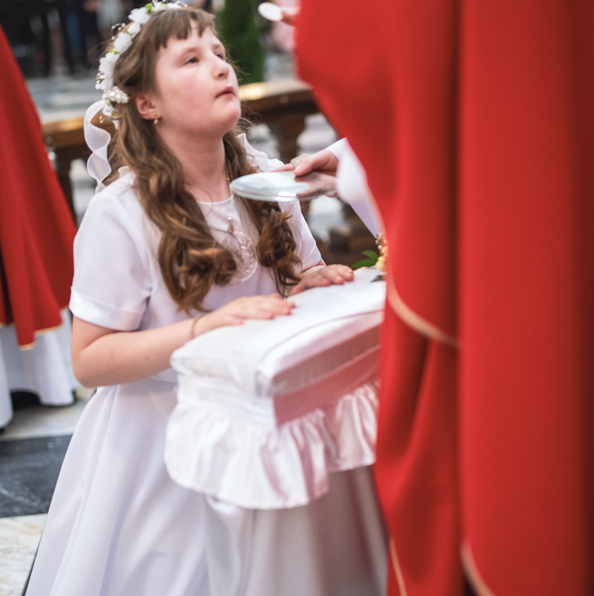 Dziewczynka w białej sukience i wianku na głowie przyjmuje komunię świętą, klęcząc na białej poduszce w kościele.