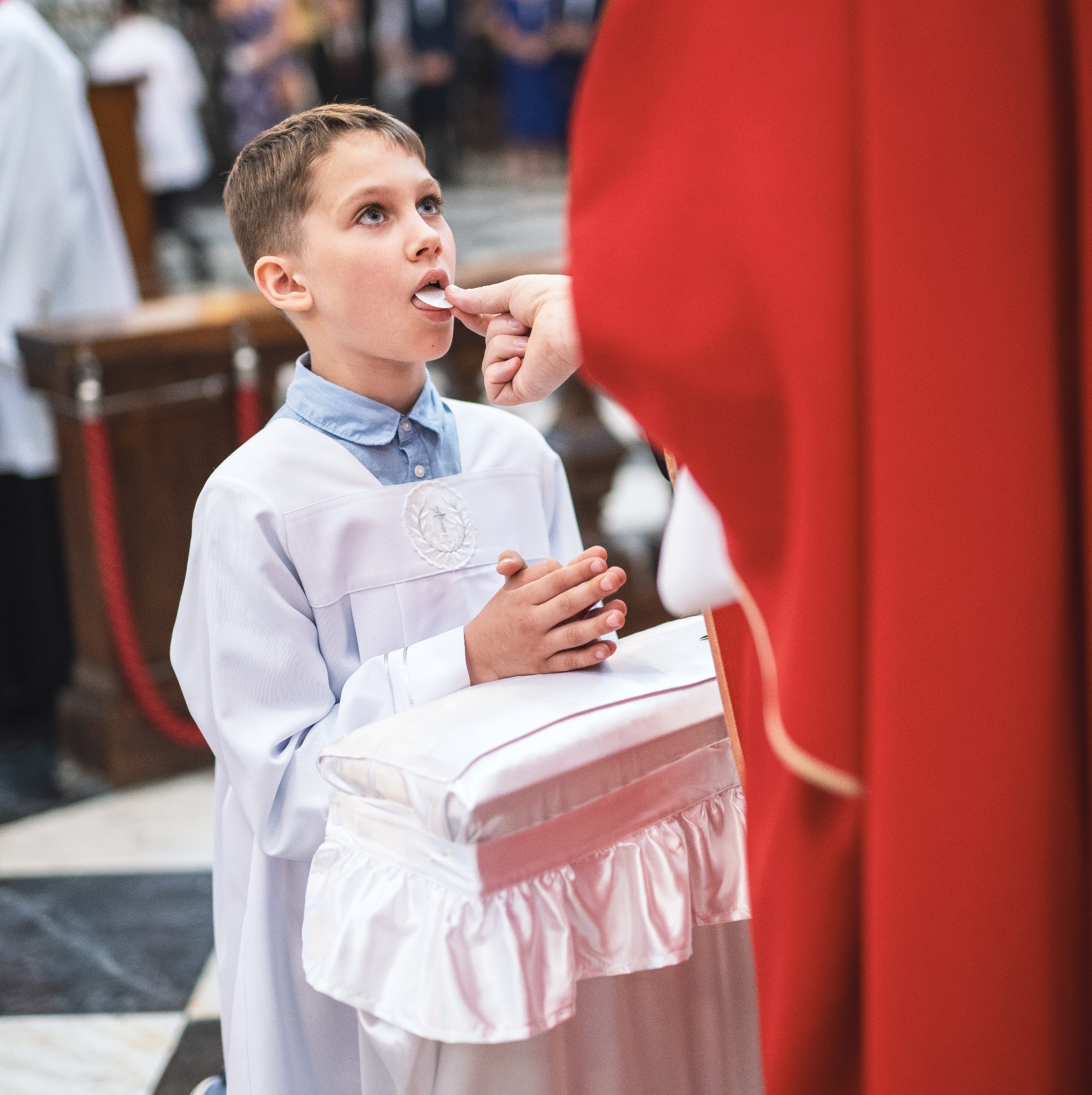 Chłopiec w albie przyjmuje komunię świętą na tle kościelnego wnętrza, z widocznym detalem haftowanego emblematu na stroju.