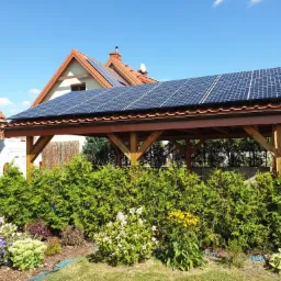 Drewniana wiata samochodowa z panelami słonecznymi na dachu, na tle domu jednorodzinnego z czerwoną dachówką i błękitnym niebem, z przodu widoczne ozdobne krzewy i kwiaty.
