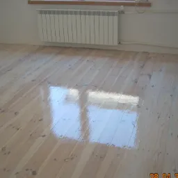 Jasny, drewniany parkiet po cyklinowaniu, z widocznym połyskiem lakieru i odbiciem okna, na tle białej ściany z grzejnikiem.