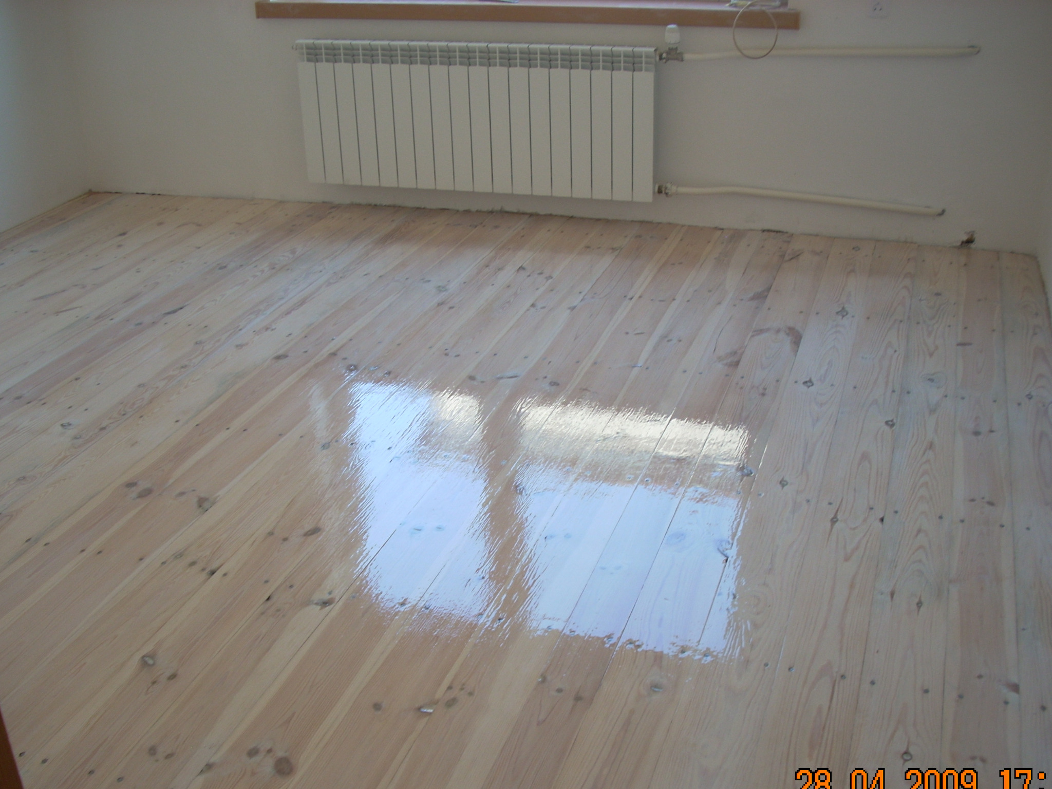 Jasny, drewniany parkiet po cyklinowaniu, z widocznym połyskiem lakieru i odbiciem okna, na tle białej ściany z grzejnikiem.