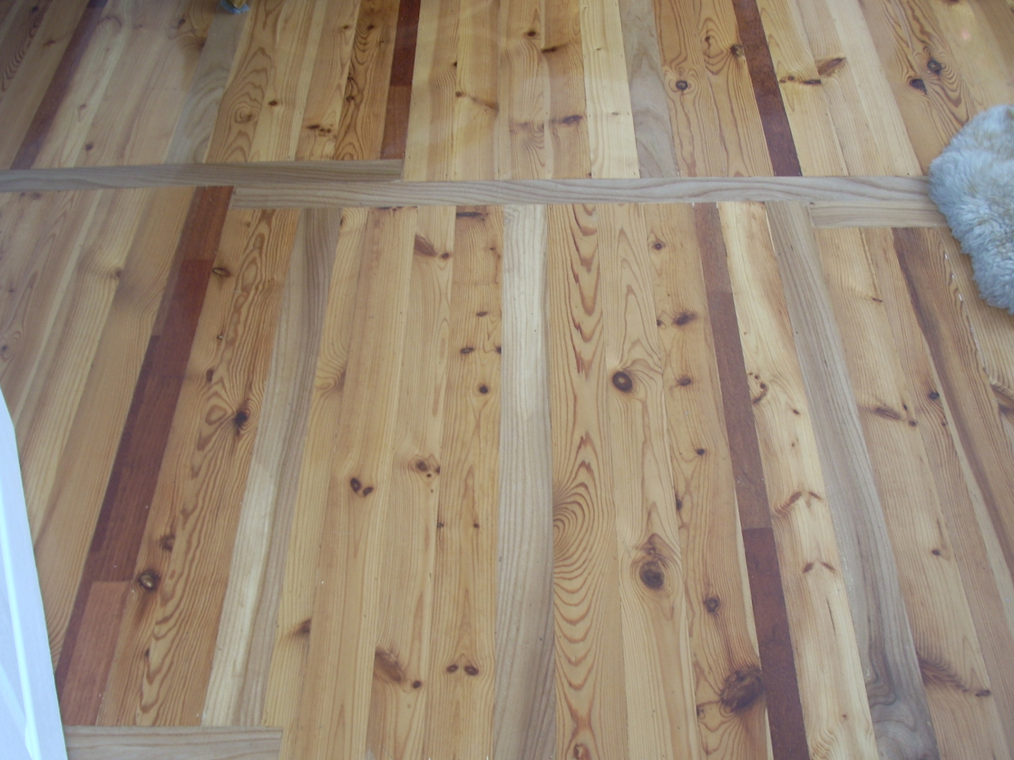 Fragment podłogi z desek z widocznymi różnicami w odcieniach drewna, ułożonych wzdłużnie z wstawkami z innego gatunku drewna, częściowo przykrytej futrzanym dywanikiem.