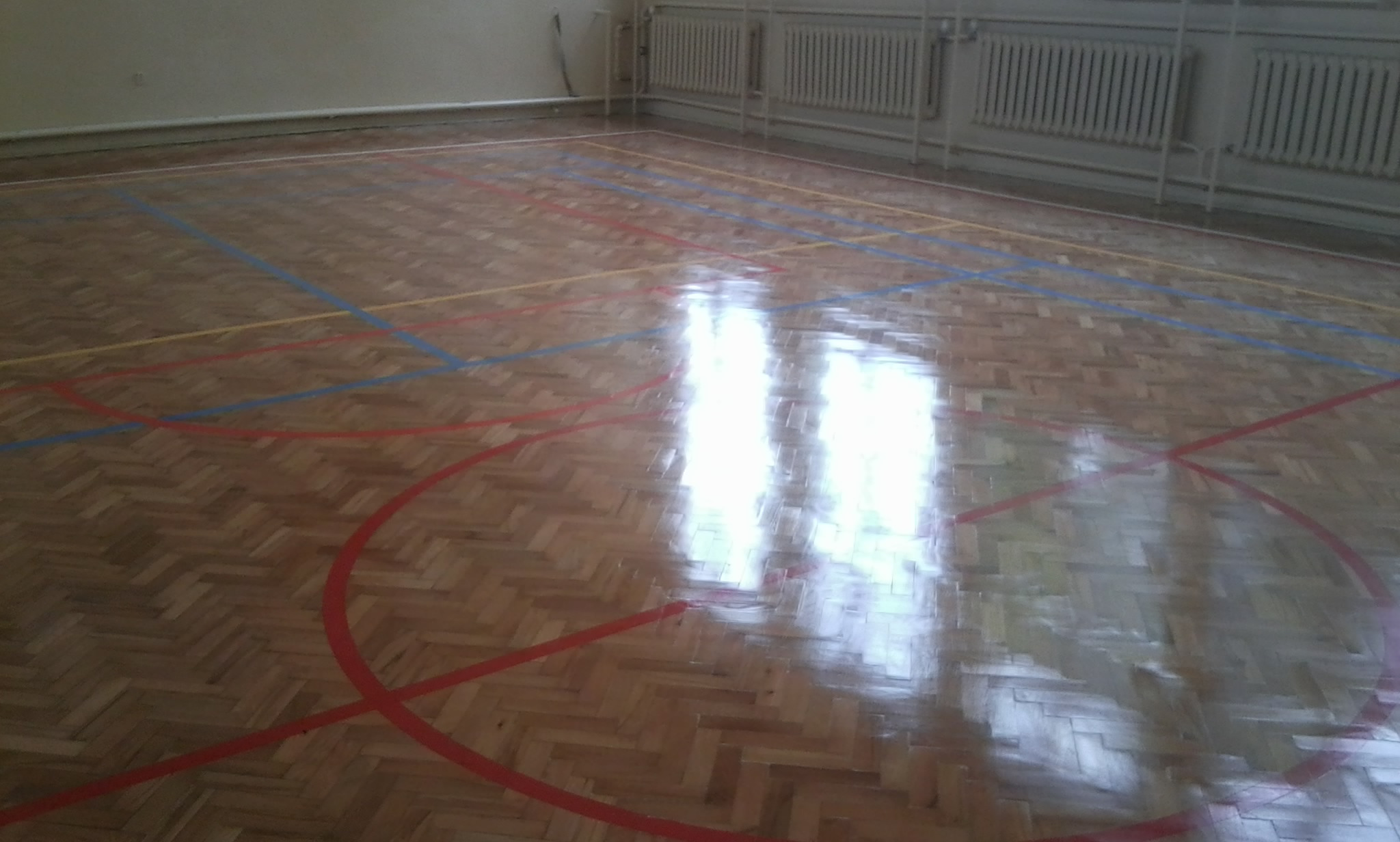 Parkiet w sali gimnastycznej z wyraźnie widocznymi liniami boiska w kolorach czerwonym, niebieskim i żółtym, odbijający światło okien. Parkiet ułożony we wzór jodełki.