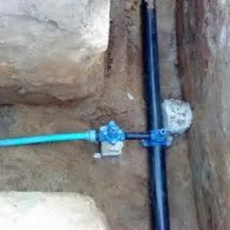 Niebieski przewód wodny podłączony do czarnej rury kanalizacyjnej w wykopie ziemnym, widoczne złącze z zaworem i obejmy mocujące.