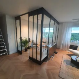 Szklana ścianka działowa z czarnymi metalowymi profilami oddzielająca przestrzeń biurową od salonu z parkietem w jodełkę i rośliną doniczkową.