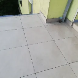 Wyłożony jasnymi płytkami taras z widocznymi krzyżykami dystansowymi przy ścianie budynku pomalowanej na żółto, z boku widoczny czarny metalowy element konstrukcyjny.