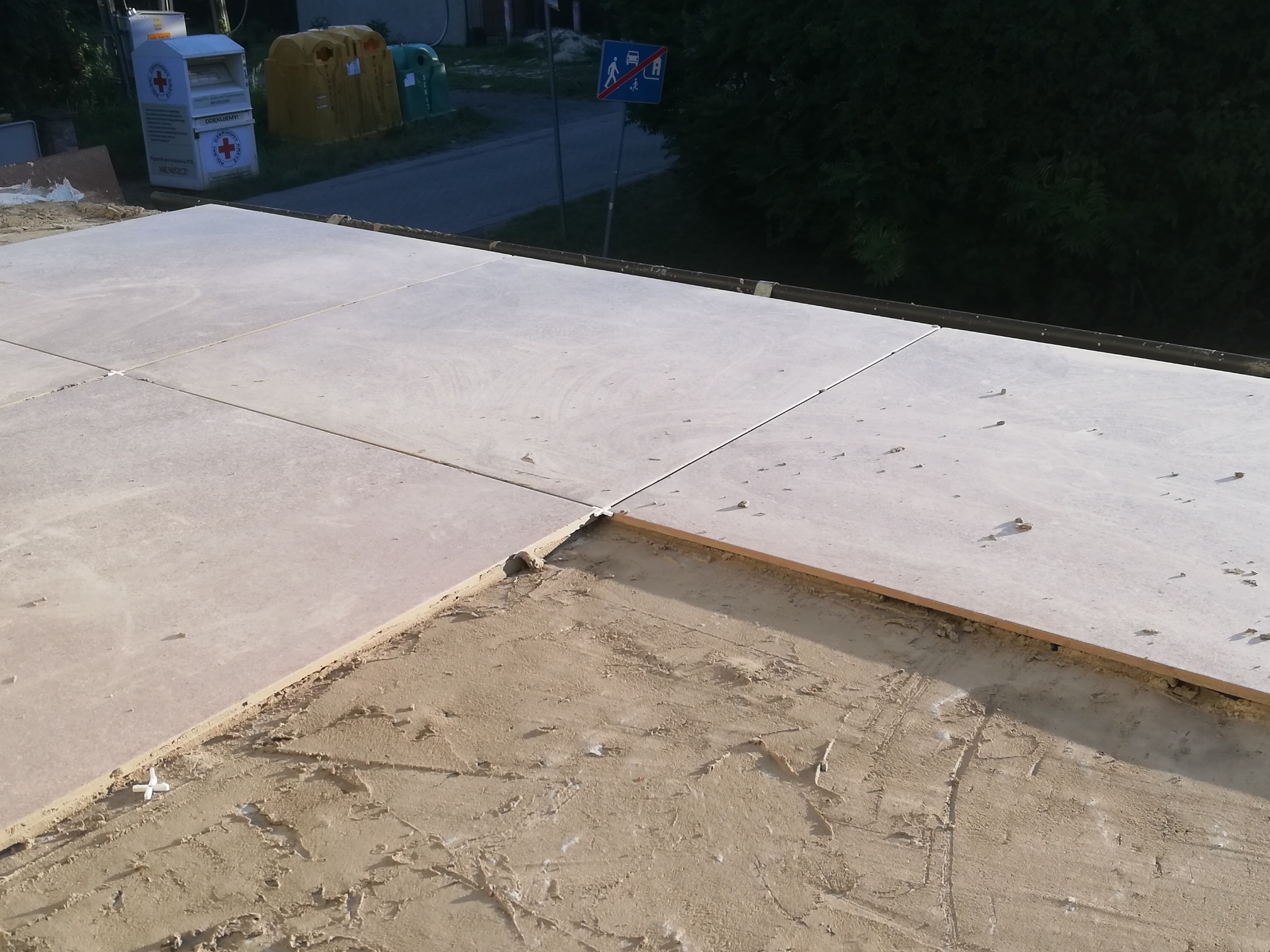 Układanie beżowych płyt na zewnątrz, widoczne krzyżyki dystansowe i podłoże z zaprawy, w tle pojemniki na odzież i śmietniki.