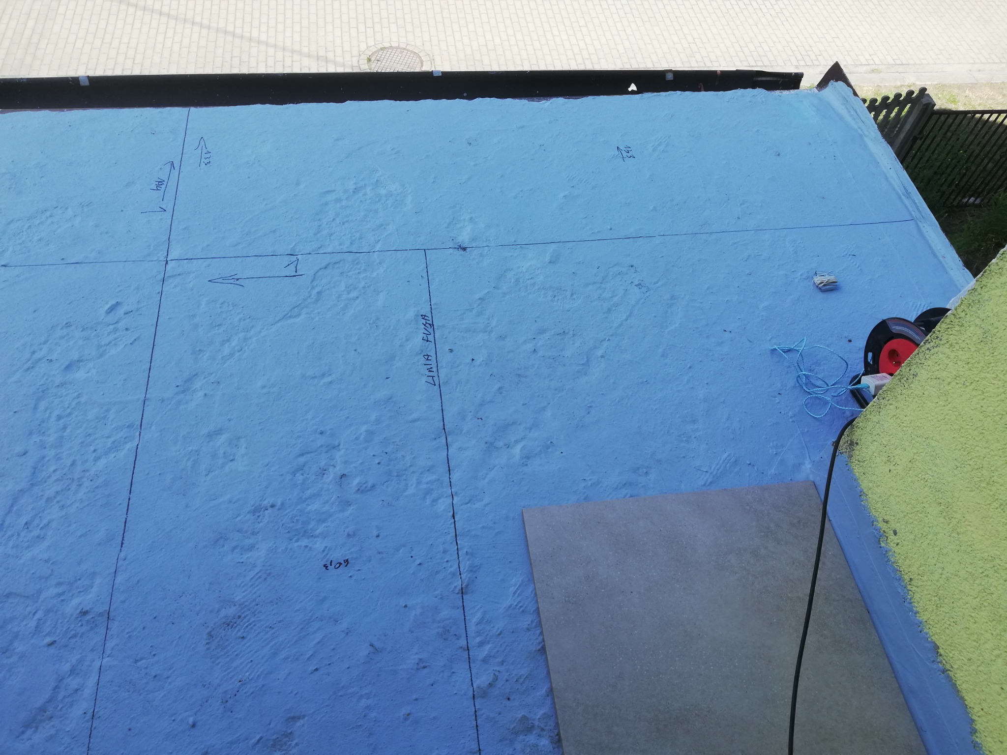 Przygotowanie podłoża pod płytki, z naniesionymi liniami i wymiarami na niebieskiej hydroizolacji, widoczna płytka i narzędzia do cięcia.