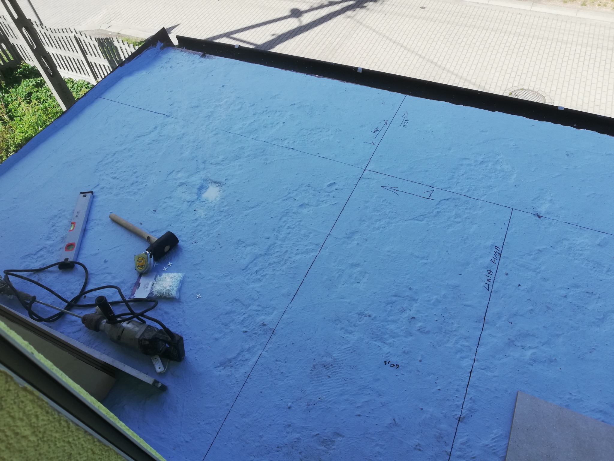 Niebieska hydroizolacja na posadzce z naniesionymi liniami pomiarowymi, poziomica, młotek gumowy, wiertarka i krzyżyki dystansowe do płytek.