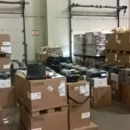 Magazyn z zapakowanymi drukarkami i laptopem na kartonach, widoczne bramy magazynowe w tle.