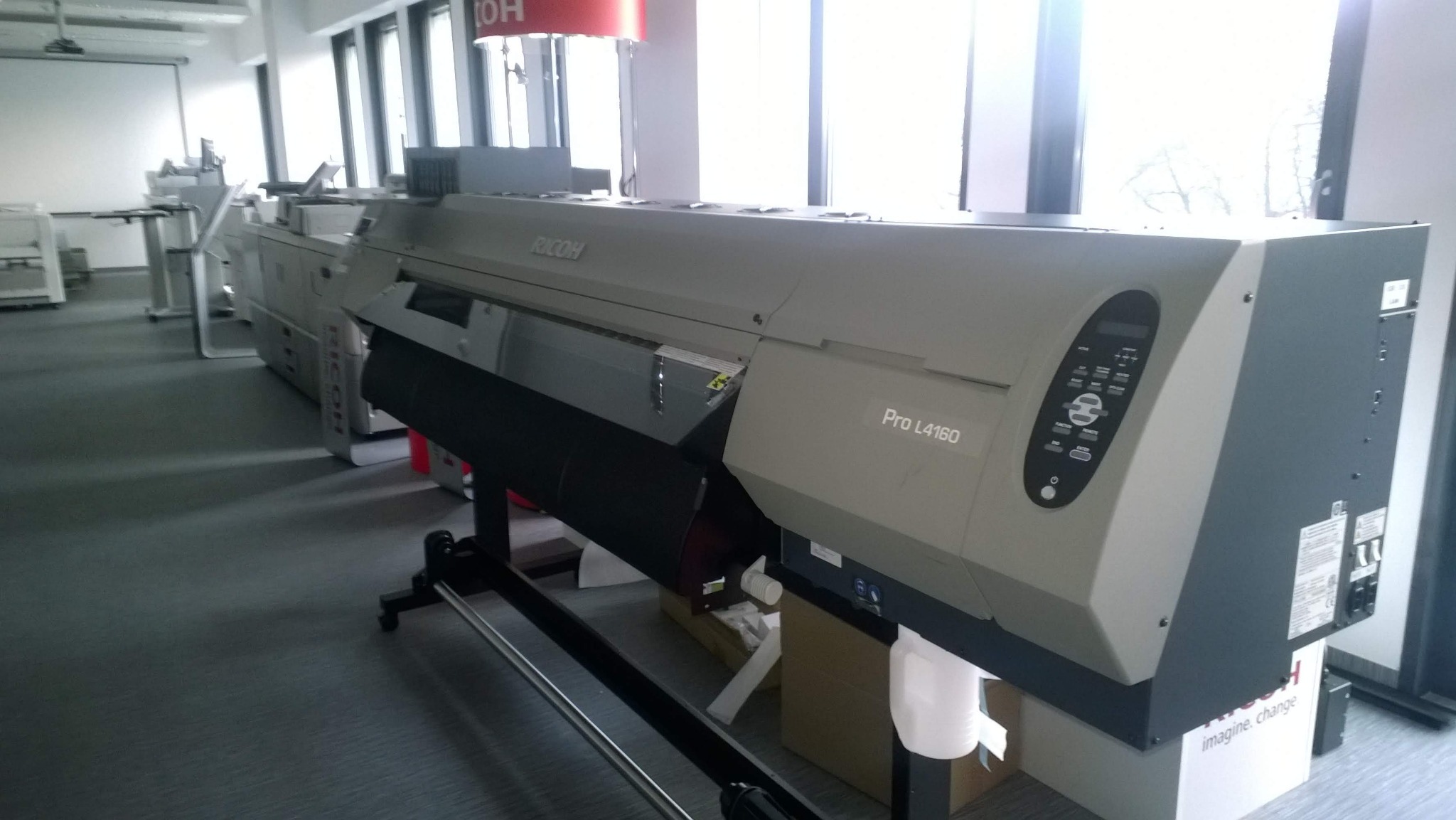 Szerokoformatowa drukarka Ricoh Pro L4160 w jasnym pomieszczeniu biurowym, widoczne inne urządzenia drukarskie w tle.