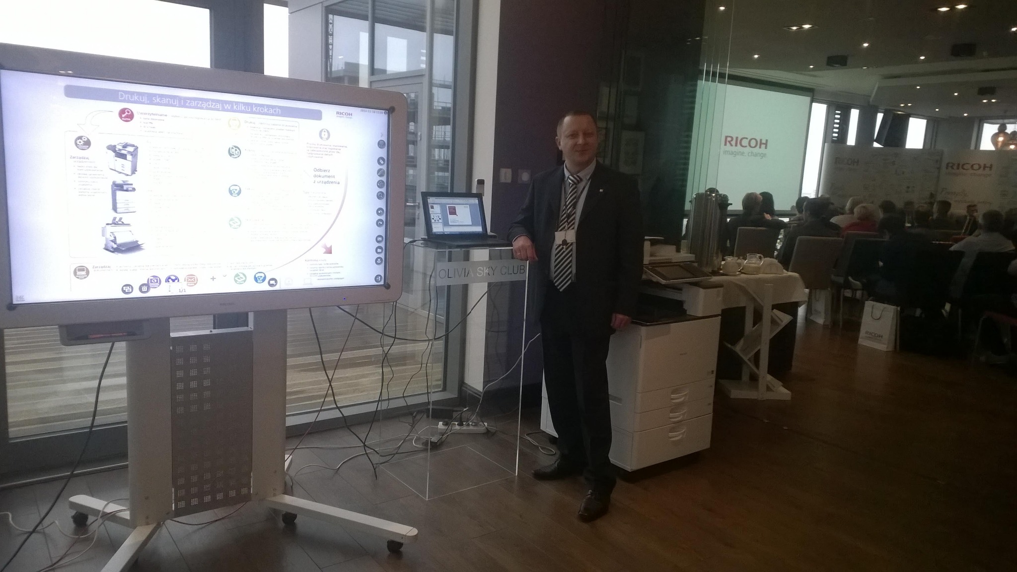 Prezentacja firmy IT na konferencji: mężczyzna w garniturze stoi obok monitora interaktywnego prezentującego funkcje drukarki Ricoh, z laptopem na przezroczystym podium i drukarką obok, w tle sala...