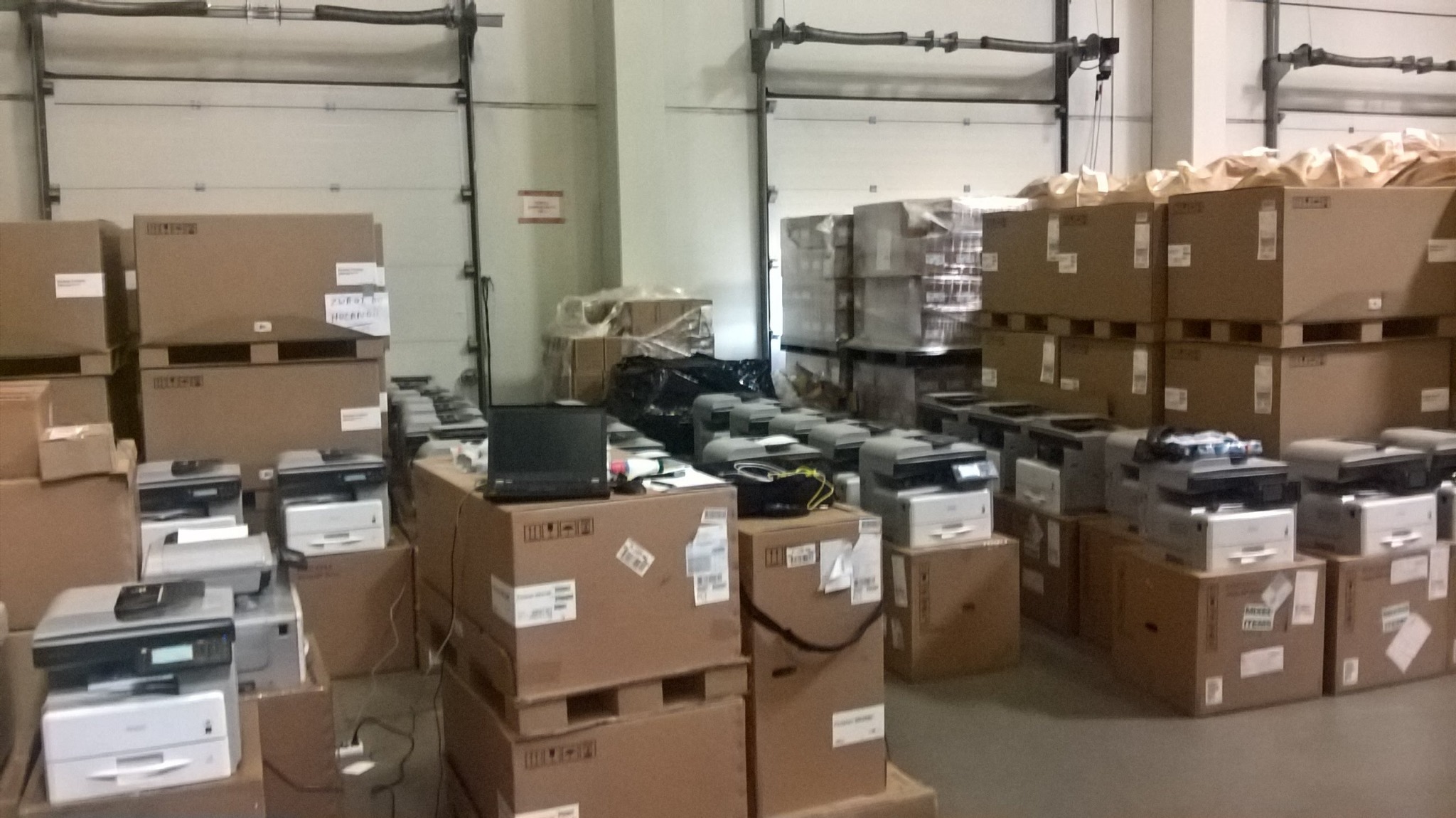 Magazyn z zapakowanymi drukarkami i laptopem na kartonach, widoczne bramy magazynowe w tle.