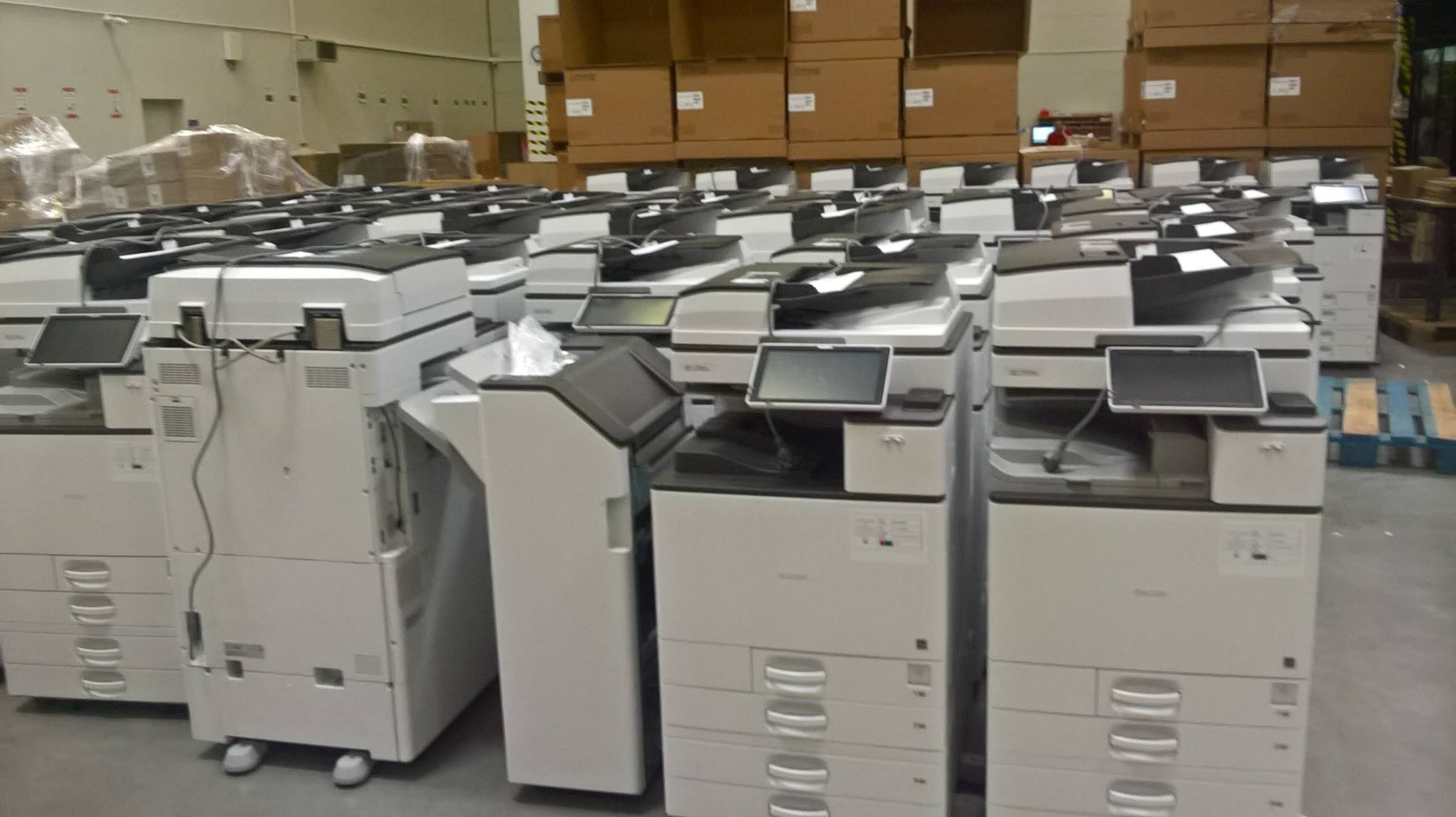 Rząd nowych, zapakowanych i gotowych do dystrybucji kserokopiarek w magazynie z kartonami w tle.