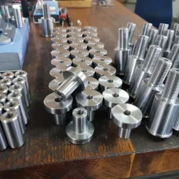 Partie metalowych elementów obrobionych maszynowo, w tym tuleje, kołnierze i gwintowane trzpienie, ułożone rzędami na drewnianym stole warsztatowym, obok suwmiarki i narzędzi.