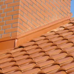 Czerwona dachówka ceramiczna na dachu domu, widoczny komin z cegły klinkierowej i obróbka blacharska w kolorze dachówki, na tle błękitnego nieba.