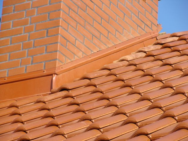 Czerwona dachówka ceramiczna na dachu domu, widoczny komin z cegły klinkierowej i obróbka blacharska w kolorze dachówki, na tle błękitnego nieba.