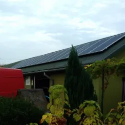 Zielony budynek z panelami słonecznymi na dachu, widoczny czerwony samochód dostawczy i roślinność w ogrodzie, pochmurne niebo.