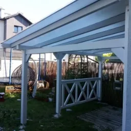 Biała pergola z przezroczystym zadaszeniem, drewnianą balustradą w kształcie krzyży, widok na ogród z huśtawką wiszącą i fragmentem domu w tle, częściowo wykończona podłoga z desek.