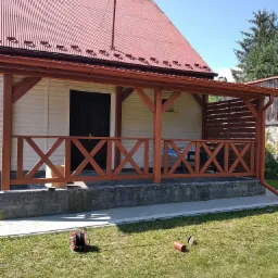 Zadaszony taras przy domu z drewnianą balustradą w kształcie krzyży, czerwony dach z rynnami, na trawniku leżą rury i zwinięty wąż ogrodowy.