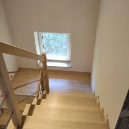 Jasne drewniane schody z metalowymi poręczami, widok z g&oacute;ry, małe okno na p&oacute;łpiętrze, fragment ściany w trakcie wykończenia.