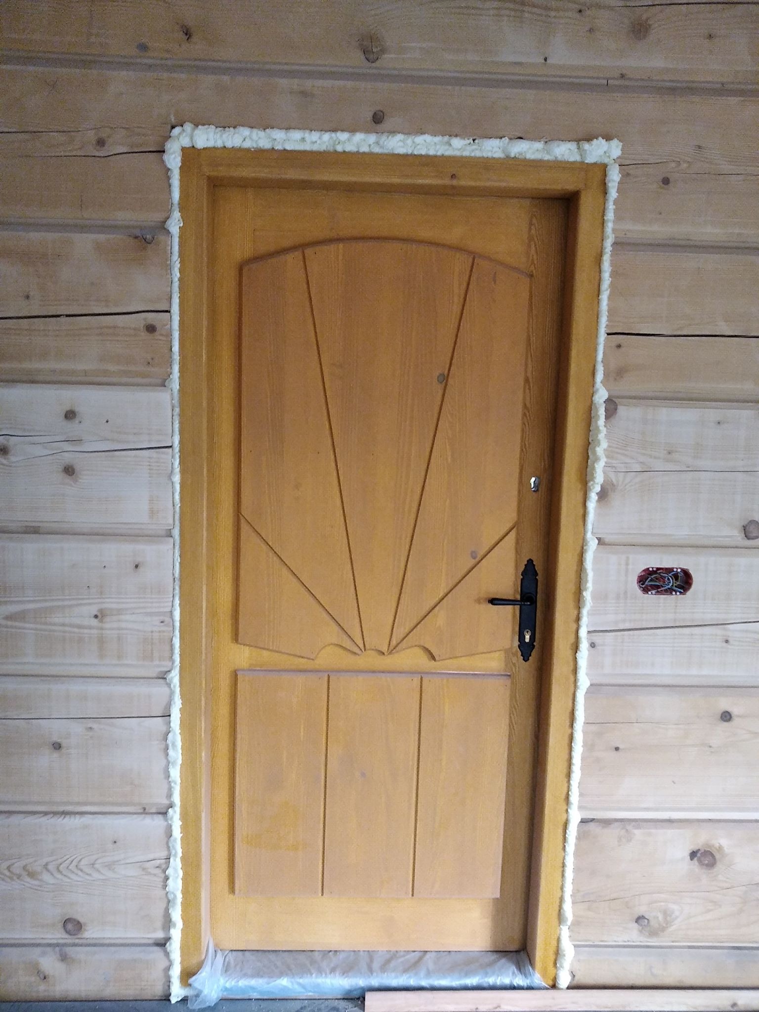 Montaż drewnianych drzwi w drewnianej ścianie, uszczelnione pianką montażową, widoczny fragment instalacji elektrycznej.