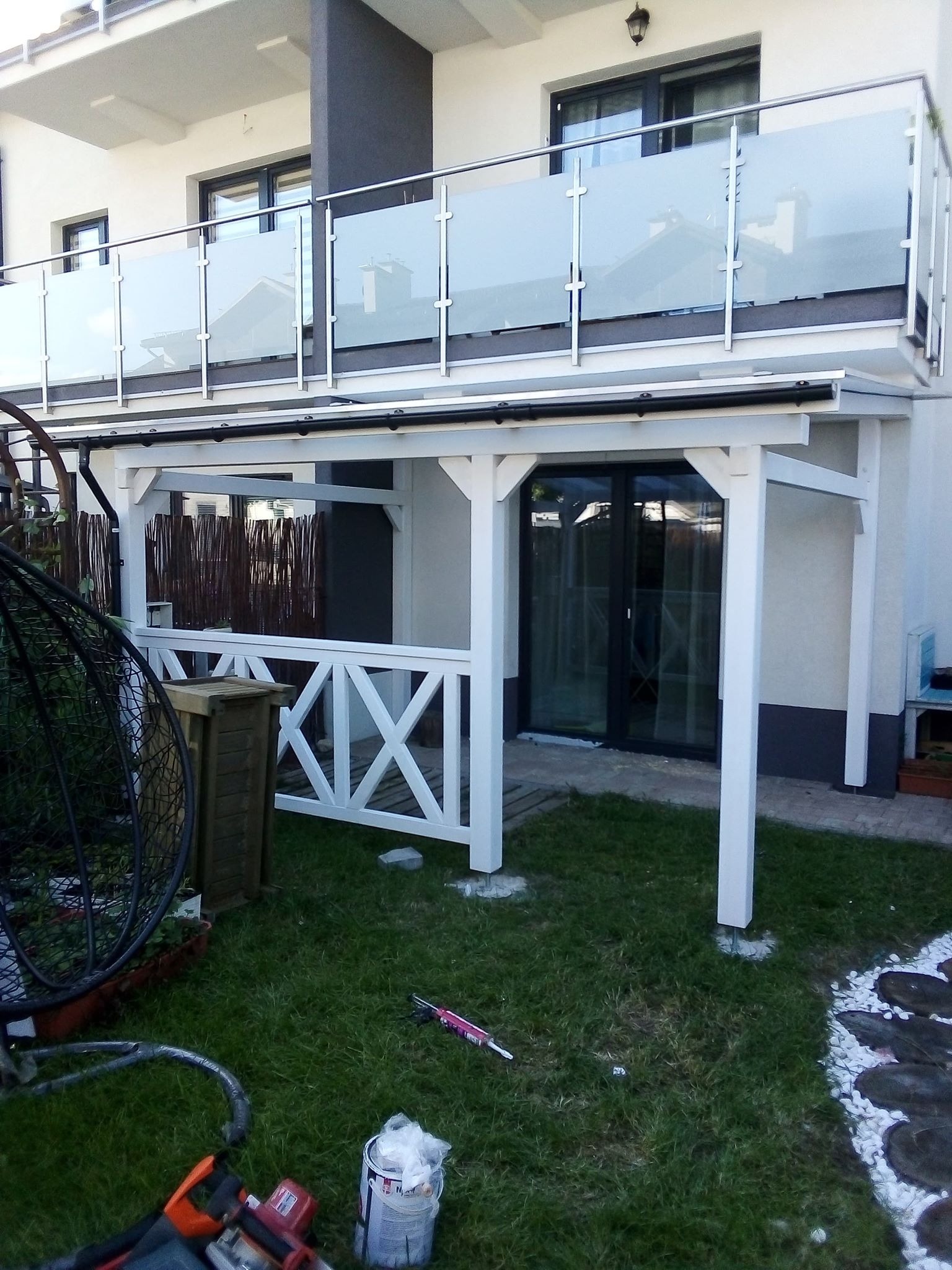 Biała pergola z drewnianą balustradą w kształcie krzyży, przylegająca do domu z balkonem z szklanymi panelami, widoczne narzędzia malarskie na trawie.