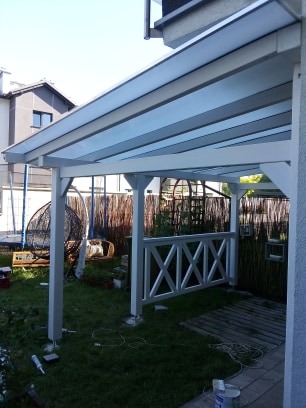 Biała pergola z przezroczystym zadaszeniem, drewnianą balustradą w kształcie krzyży, widok na ogród z huśtawką wiszącą i fragmentem domu w tle, częściowo wykończona podłoga z desek.