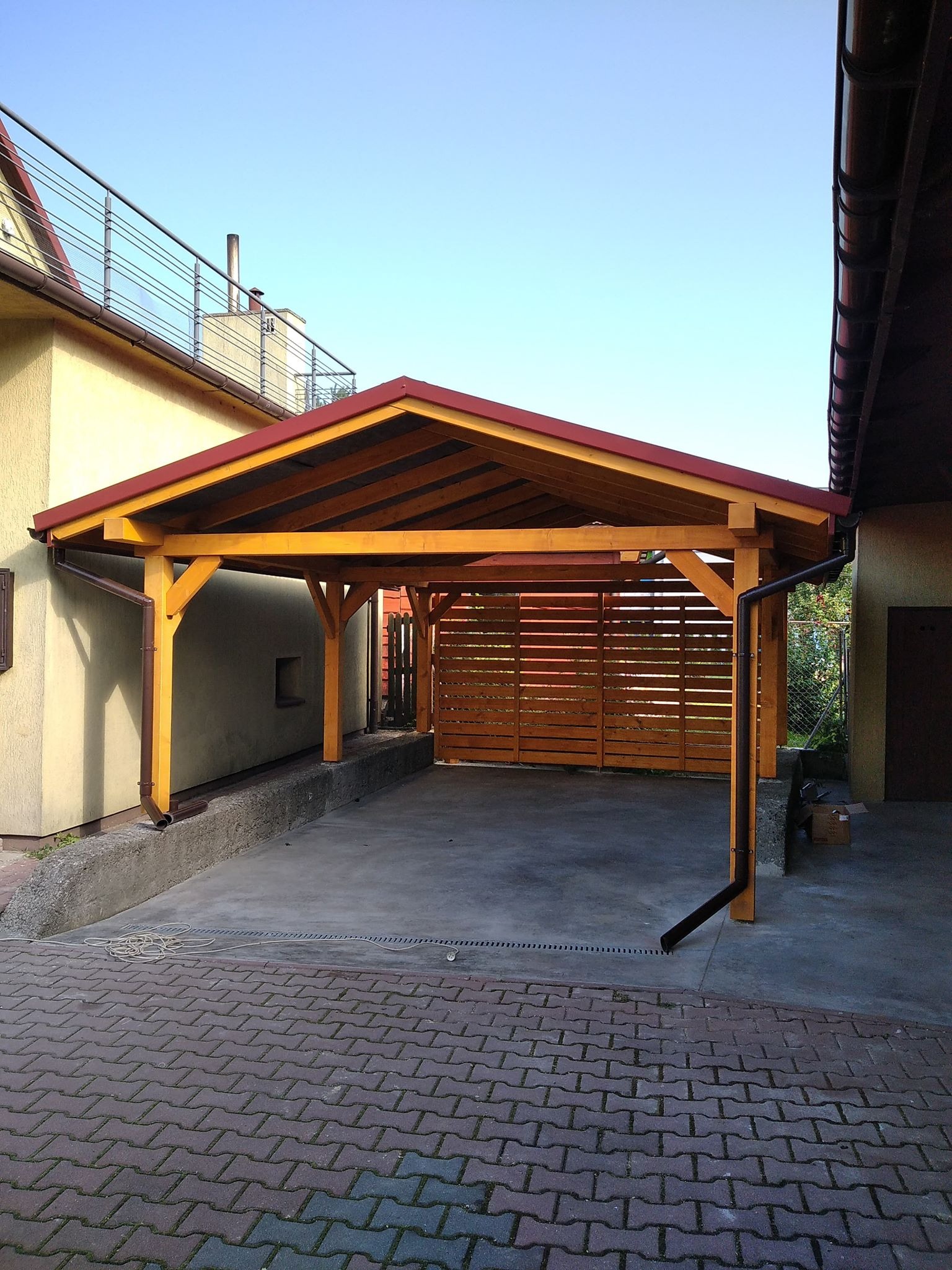Drewniana wiata garażowa z czerwonym dachem, przylegająca do budynku, z widoczną drewnianą ażurową ścianą z tyłu, na betonowej posadzce i kostce brukowej.
