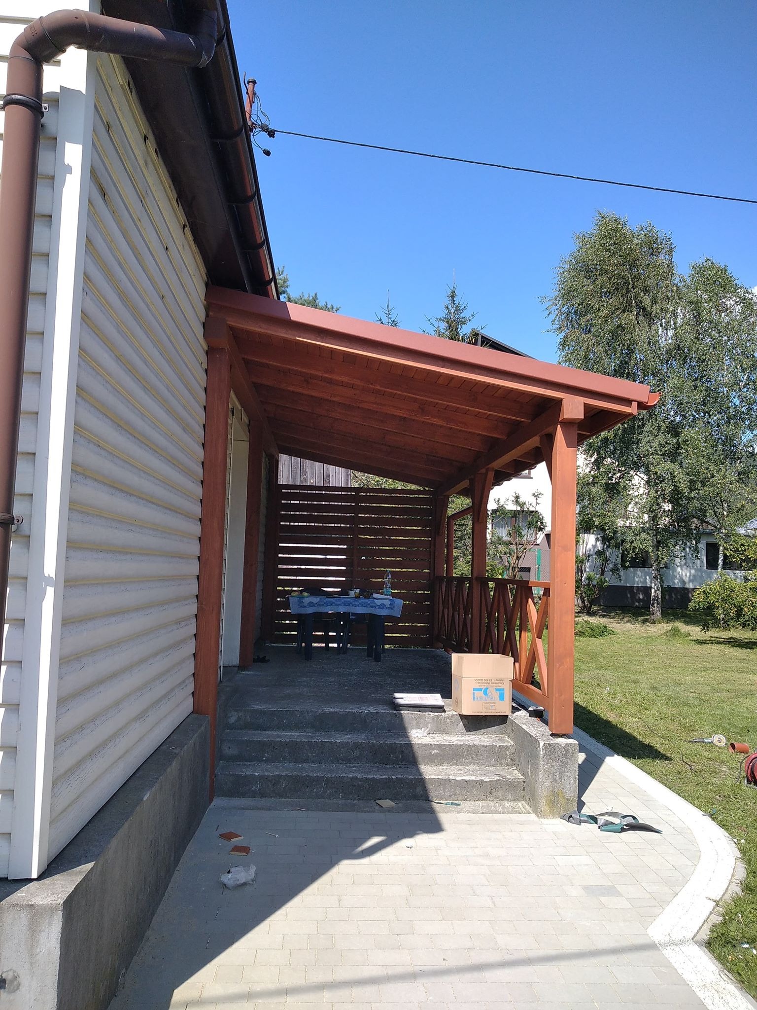 Drewniana pergola przylegająca do budynku z sidingiem, z widocznymi schodami wejściowymi i częściowo zadaszonym tarasem, na którym stoi stół nakryty niebieskim obrusem.
